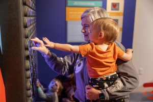 An adult helps a small child touch a large interactive light board. Other children play nearby in a colorful, indoor museum setting. *AI generated alt text
