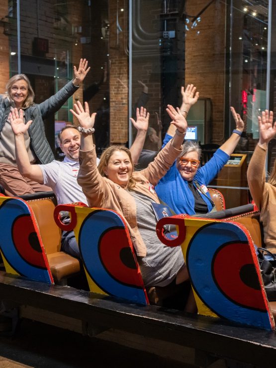 Seven adults sit in a colorful roller coaster car inside a building, all raising their hands in the air as if on a ride. The background includes glass walls, lighting fixtures, and exposed brick within the museum. *AI generated alt text
