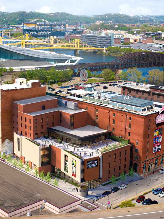 Aerial view of downtown Pittsburgh buildings, with bridges and a river in the background. A rooftop terrace can be seen, and several cars are parked along the nearby streets, illustrating the urban landscape surrounding the museum. *AI generated alt text