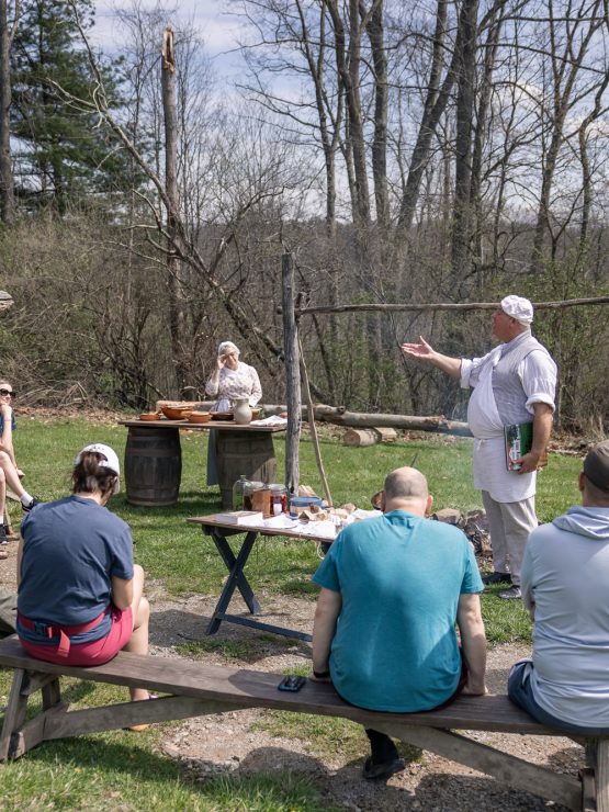 A group of visitors sits outdoors on benches, listening to an interpreter dressed in historical clothing speak near a log cabin. Another staff member stands behind a table with supplies. The scene is set among trees and grass at the museum. *AI generated alt text