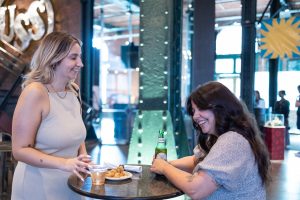 Two women are seated at a high-top table with drinks and snacks in a bright space featuring industrial architectural elements. *AI generated alt text