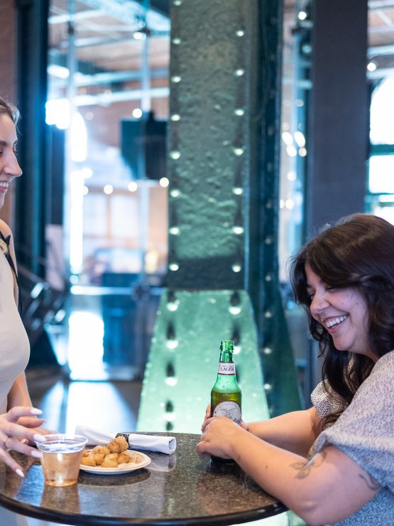 Two women are seated at a high-top table with drinks and snacks in a bright space featuring industrial architectural elements. *AI generated alt text