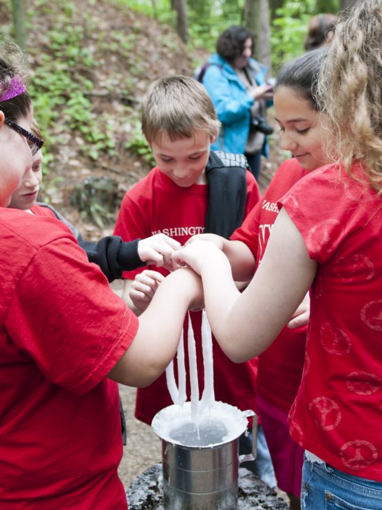 Four children wearing red shirts operate a hand-crank ice cream machine outdoors during a second Saturday event at the museum. Trees and additional visitors appear in the background. *AI generated alt text