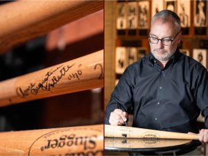 A man wearing glasses signs a wooden Louisville Slugger baseball bat. A close-up reveals the pen marking the bat, with sports memorabilia and historic photographs displayed in the background of the museum exhibit. *AI generated alt text