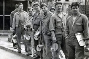 A group of men in historical military uniforms stand in a line outdoors, holding metal trays and mugs. They appear relaxed near a fence and a building in the background. *AI generated alt text