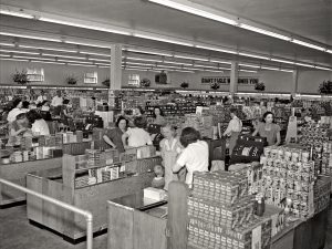 Black and white image of a mid-20th century supermarket, with shoppers, cashiers at checkout lanes, and shelves of canned goods. *AI generated alt text