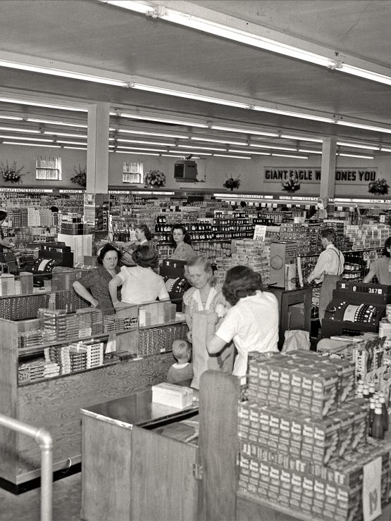 Black and white image of a mid-20th century supermarket, with shoppers, cashiers at checkout lanes, and shelves of canned goods. *AI generated alt text