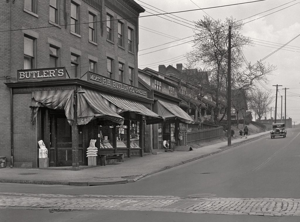 Black-and-white photo showing Butler’s store with awnings and goods outside, shops along street, vintage car, houses, and trees. *AI generated alt text