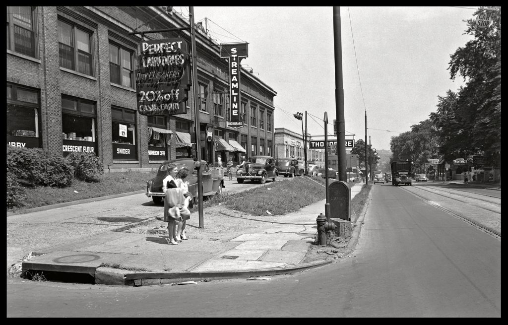 Historical street scene with two children, vintage cars, shopfronts, trees, overhead wires, and a passing streetcar under clear skies. *AI generated alt text