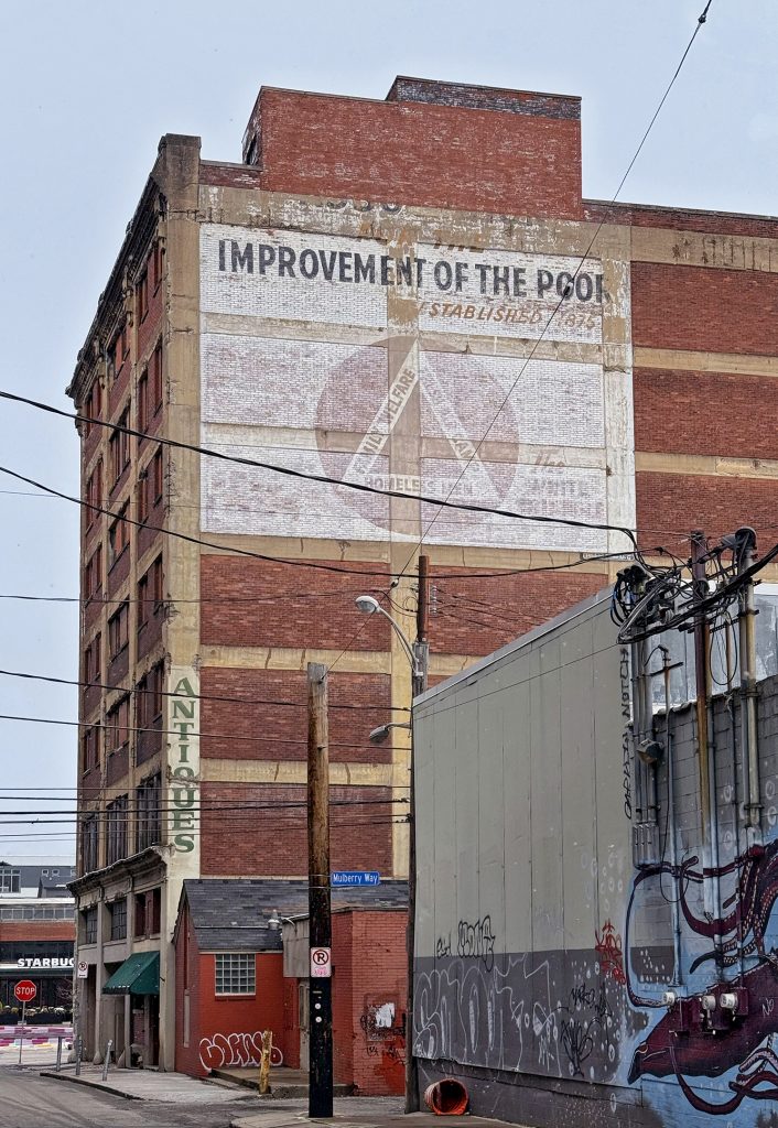 Historic mural on brick building reads IMPROVEMENT OF THE POOR above faded text; antique shop, street art, and urban features nearby. *AI generated alt text