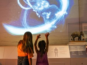 Two children with arms raised face a glowing mammoth projection; nearby are a model mammoth and informative museum displays. *AI generated alt text