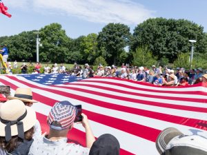A large group of people display a giant American flag outdoors on a sunny day during civic season, with several individuals wearing hats and some taking photographs; trees and clear blue sky are visible in the background. *AI generated alt text