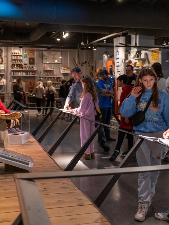 A group of children and adults participate in a field trip, interacting with hands-on exhibits and reading informational displays in a crowded history museum. Some visitors take notes while a mannequin is positioned on a porch scene exhibit. *AI generated alt text