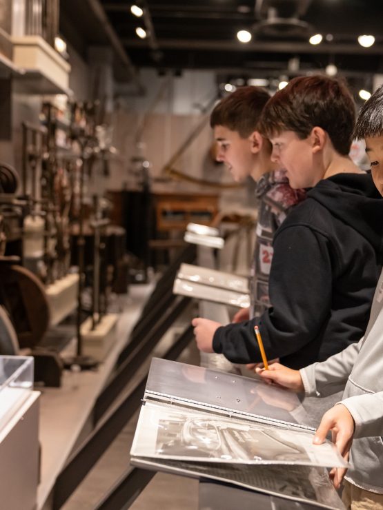 Three boys stand at display panels in a history museum, observing exhibits and writing notes. Artifacts, such as a uniform, are displayed in glass cases and on the walls behind them. *AI generated alt text