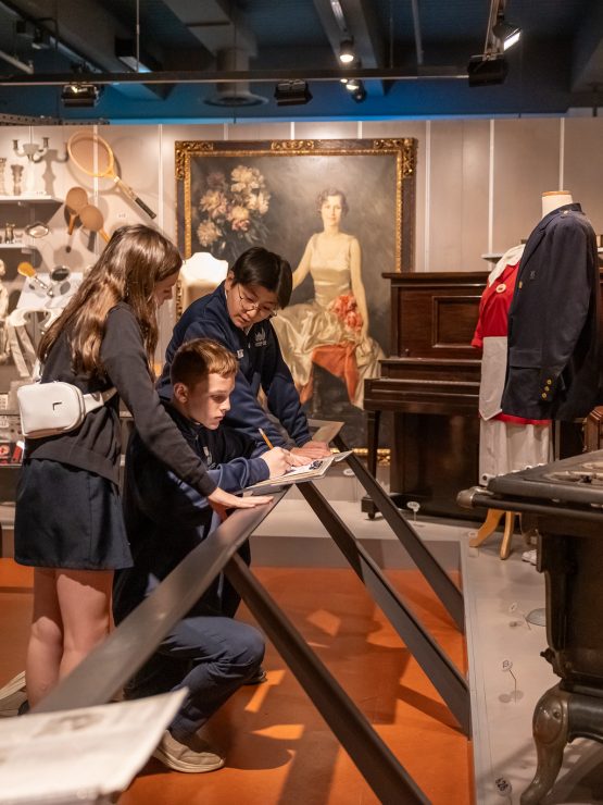 Three visitors on a field trip observe exhibits and read informational displays in a museum room featuring historical artifacts such as a stove, vintage clothing, a piano, and a large portrait of a woman hanging on the wall. *AI generated alt text