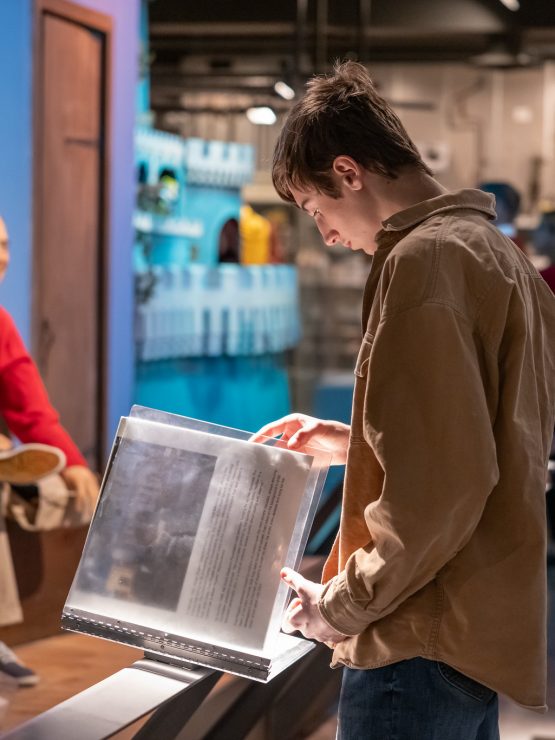 A young man stands reading an exhibit display in a history museum. In the background, a wax figure of a man dressed in a red sweater is visible, along with other visitors observing various exhibits. *AI generated alt text