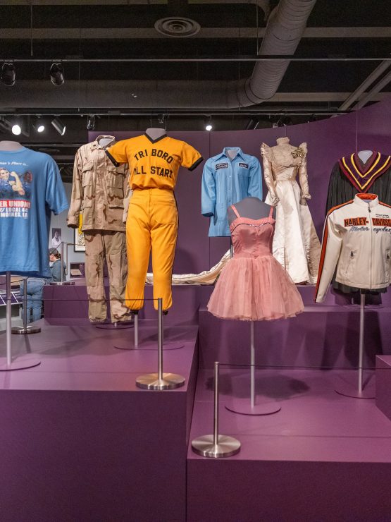 A museum exhibit displays an array of colorful costumes, uniforms, and dresses arranged on mannequins and stands. The setting features purple walls and platforms. A football helmet is positioned on the lower right. *AI generated alt text