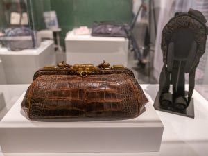A vintage brown leather handbag with a metal clasp is displayed in a museum exhibit, surrounded by other historical artifacts and objects in the background. *AI generated alt text