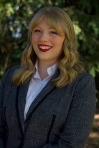 A woman with blond wavy hair, dressed in a gray blazer over a white shirt, stands smiling in front of a backdrop of green trees. *AI generated alt text