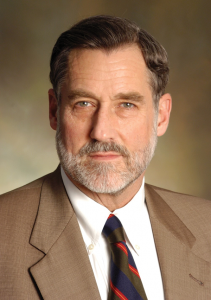 A middle-aged man with a gray beard and neatly combed hair, dressed in a brown suit, white shirt, and striped tie, looks directly at the camera. He is positioned against a softly blurred, neutral background in a formal portrait style. *AI generated alt text