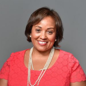 A woman with short brown hair, wearing a coral lace top, pearl earrings, and a double-strand pearl necklace, smiles while posing against a plain gray background in the style of traditional portrait photography. *AI generated alt text
