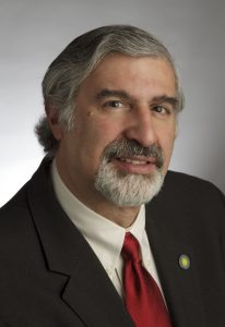 A middle-aged man with gray hair and a beard, dressed in a dark suit, white shirt, and red tie, is pictured in a professional headshot against a neutral background. The photographic style emulates formal portraiture commonly found in museum collections. *AI generated alt text