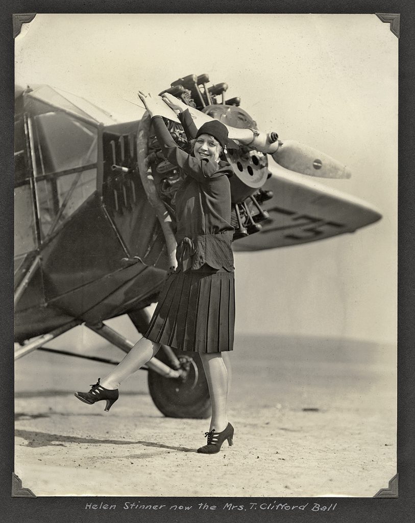 A woman in a cloche hat and pleated dress stands with arms raised beside an early airplane, with the aircraft’s propeller and engine components clearly visible in the background. *AI generated alt text