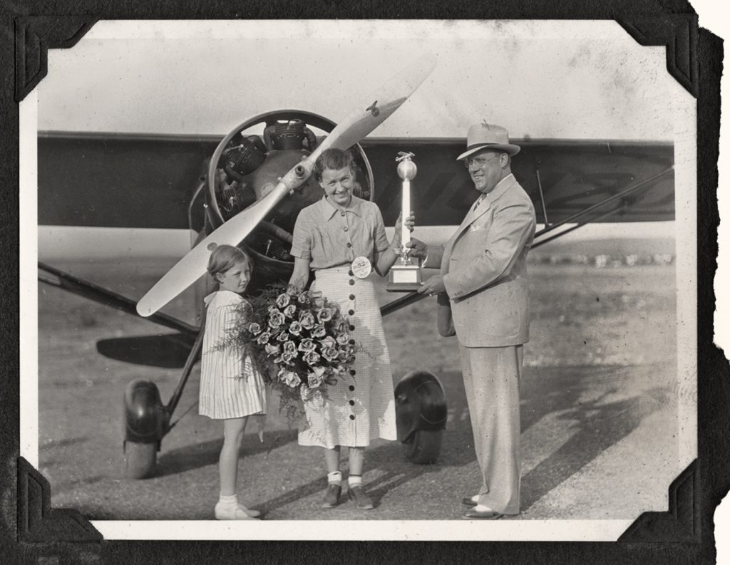 A historical photograph shows a woman standing in front of an airplane, smiling, while holding a medal and flowers. Beside her, a girl holds a bouquet. A man in a suit presents the woman with a trophy. The group is positioned outdoors on an airfield. *AI generated alt text