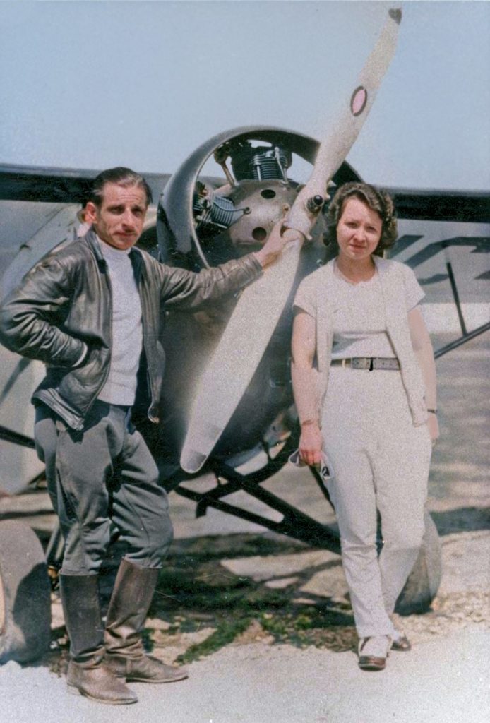 A man wearing a leather jacket and boots stands beside a woman dressed in a light-colored outfit next to a propeller airplane. The pair are outdoors in daylight, positioned near the aircraft. *AI generated alt text