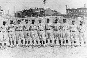 A vintage black-and-white photograph shows eleven baseball players in identical uniforms standing in a straight line on a dirt field. In the background, there are several buildings and utility poles, capturing an early 20th-century American baseball scene. *AI generated alt text