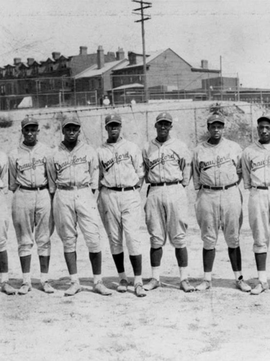 A vintage black-and-white photograph shows eleven baseball players in identical uniforms standing in a straight line on a dirt field. In the background, there are several buildings and utility poles, capturing an early 20th-century American baseball scene. *AI generated alt text