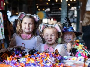 Three children in costumes, two dressed as mice with pink bows and one as a witch, are shown smiling and holding lollipops at a decorated Halloween table displaying an assortment of colorful candies and festive decorations. The setting is indoors with a blurred background. *AI generated alt text
