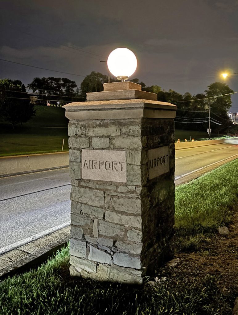 A stone pillar featuring a luminous round light on top is positioned beside a roadway at night. The word AIRPORT is engraved on two sides of the pillar. In the background, there are patches of grass, several trees, and illuminated streetlights. *AI generated alt text