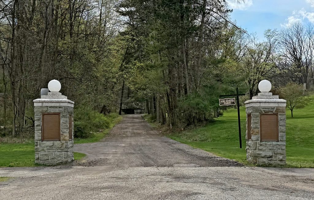 A gravel road lined with trees passes between two stone pillars, each topped with a white globe. To the right, a wooden sign displays the word “Millstone.” The surroundings are lush and green, showing early spring foliage. *AI generated alt text