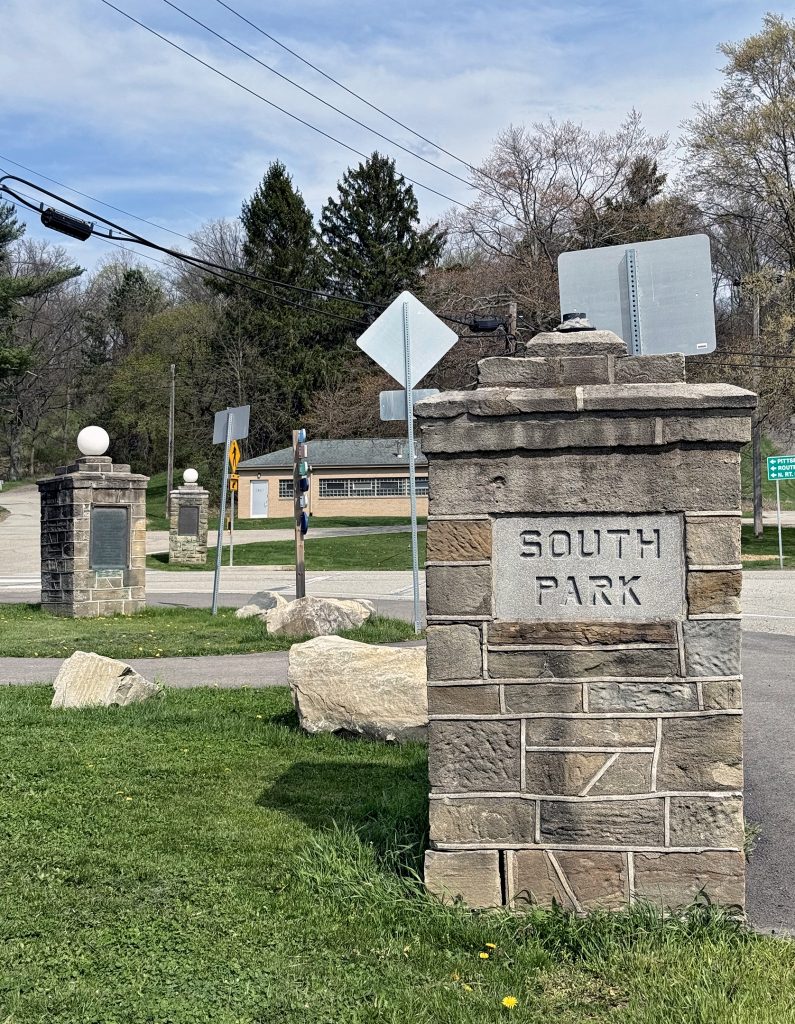 Stone pillars with SOUTH PARK engraved mark the entrance to a park. In the background, there are informational signs and a building, surrounded by green grass and trees under a clear sky. *AI generated alt text