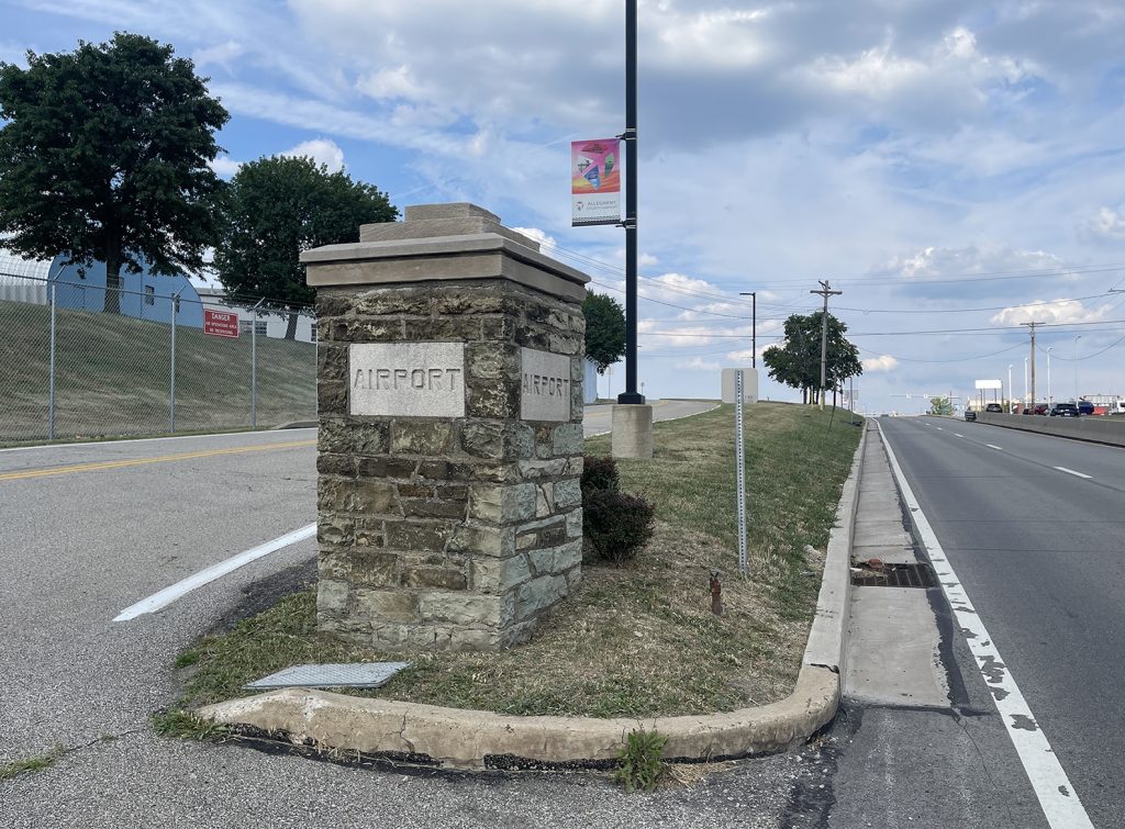 A stone pillar labeled MUSEUM stands by a roadside bordered by grass, trees, and a fence. A street lamp with a colorful banner is nearby, and the background features a partly cloudy sky. *AI generated alt text