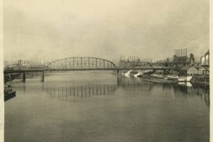 Black-and-white photograph depicting a steel bridge spanning the Monongahela River. Factories with smokestacks and several boats are visible along both riverbanks. The caption beneath the image reads: General view from Monongahela River, 1914. *AI generated alt text
