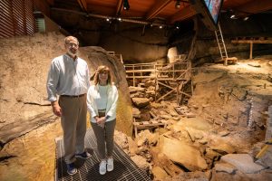 Two visitors stand on a metal platform inside the Meadowcroft indoor archaeological site, with surrounding rocks, fossils, and wooden scaffolding visible in the background under warm lighting. *AI generated alt text