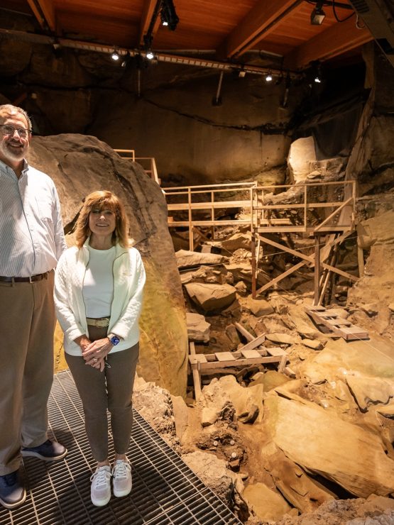 Two visitors stand on a metal platform inside the Meadowcroft indoor archaeological site, with surrounding rocks, fossils, and wooden scaffolding visible in the background under warm lighting. *AI generated alt text