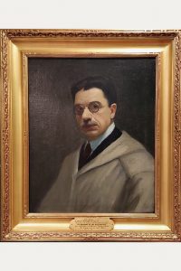 Painted portrait of A.H. Gorson shows a man with short dark hair, mustache, and glasses wearing a light-colored coat. He faces forward against a dark background. The artwork is displayed in an ornate gold frame. *AI generated alt text