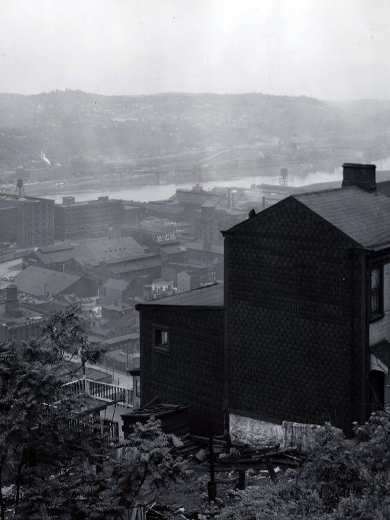 A black-and-white photograph of a hillside house overlooking an industrial cityscape featuring factories, multiple smokestacks emitting smoke, a river running through the scene, and distant hills. The background is partially obscured by mist and industrial smoke. *AI generated alt text