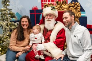 A family with a baby is seated alongside a person dressed as Santa Claus on a golden chair at Planet Ice. The group is surrounded by festive holiday decorations, including a Christmas tree and wrapped gifts. *AI generated alt text