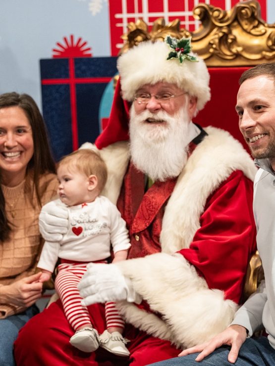 A family with a baby is seated alongside a person dressed as Santa Claus on a golden chair at Planet Ice. The group is surrounded by festive holiday decorations, including a Christmas tree and wrapped gifts. *AI generated alt text