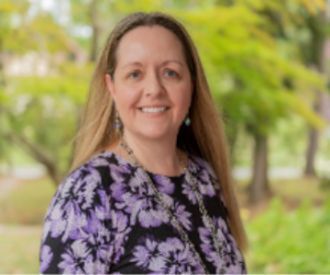 A woman with long light brown hair, wearing a purple floral top and earrings, is smiling outdoors. Green trees are blurred in the background. *AI generated alt text