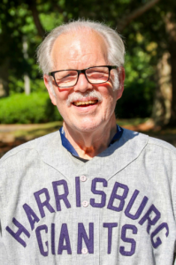 Richard Kurin, an older man with glasses and a white mustache, stands outdoors wearing a gray Harrisburg Giants baseball jersey. Trees and greenery are visible in the background. *AI generated alt text