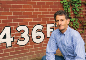 Richard Kurin, a man with short curly hair and a mustache, wearing a blue striped shirt, stands in front of a red brick wall with “436F” painted on it. Green ivy is visible at the top right corner of the wall. *AI generated alt text
