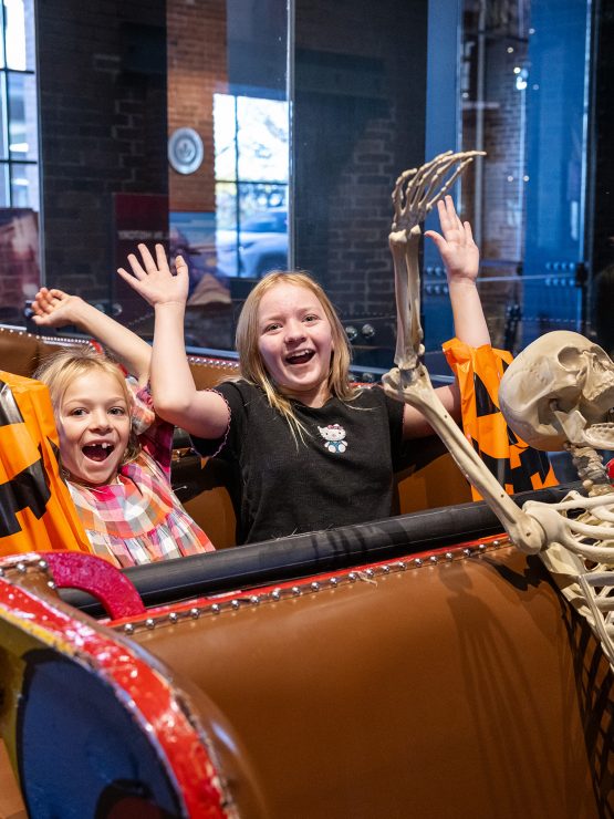 Two smiling girls and a skeleton with raised arms sit together on a roller coaster ride display indoors at the museum, surrounded by festive decorations. *AI generated alt text