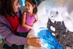 A woman and a smiling child look into an interactive museum exhibit featuring a blue water display set within a snowy landscape. *AI generated alt text