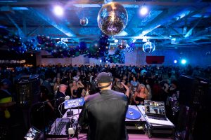 A DJ performs on stage at a museum event, overlooking a ballroom filled with disco balls and clusters of blue and silver balloons. *AI generated alt text