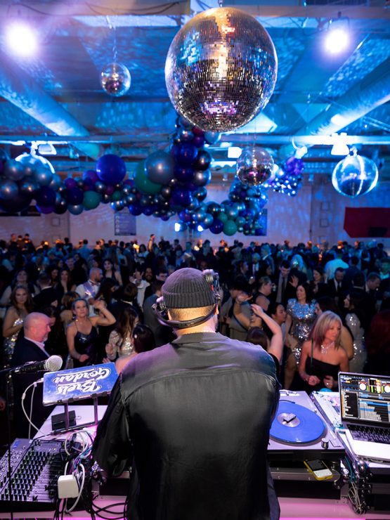 A DJ performs on stage at a museum event, overlooking a ballroom filled with disco balls and clusters of blue and silver balloons. *AI generated alt text